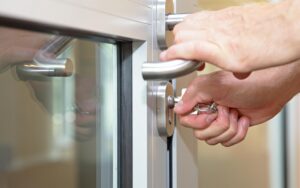 Close-up image of a person's hands locking or unlocking a glass door with a metal frame. One hand holds the door handle while the other hand inserts a key into the lock.