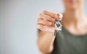 Person holding house-shaped keychain and keys, with blurred background